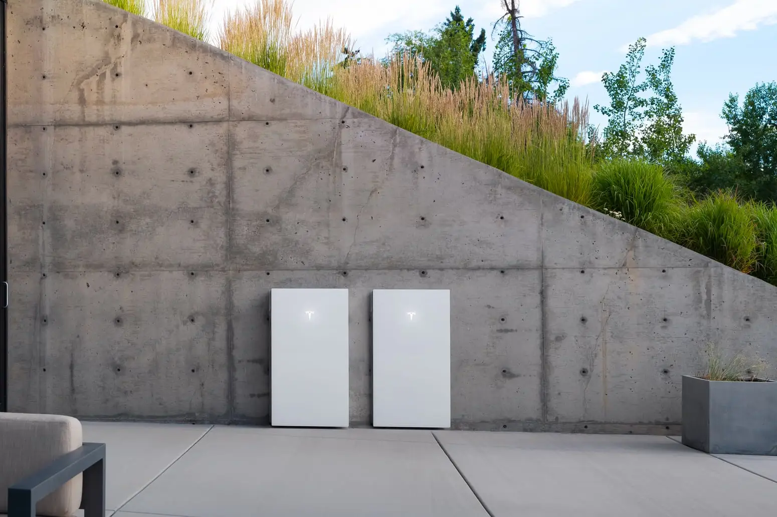 Two white Tesla-badged batteries standing side by side against a board-formed concrete retaining wall on a paved terrace, with ornamental grasses along the top of the wall and trees behind