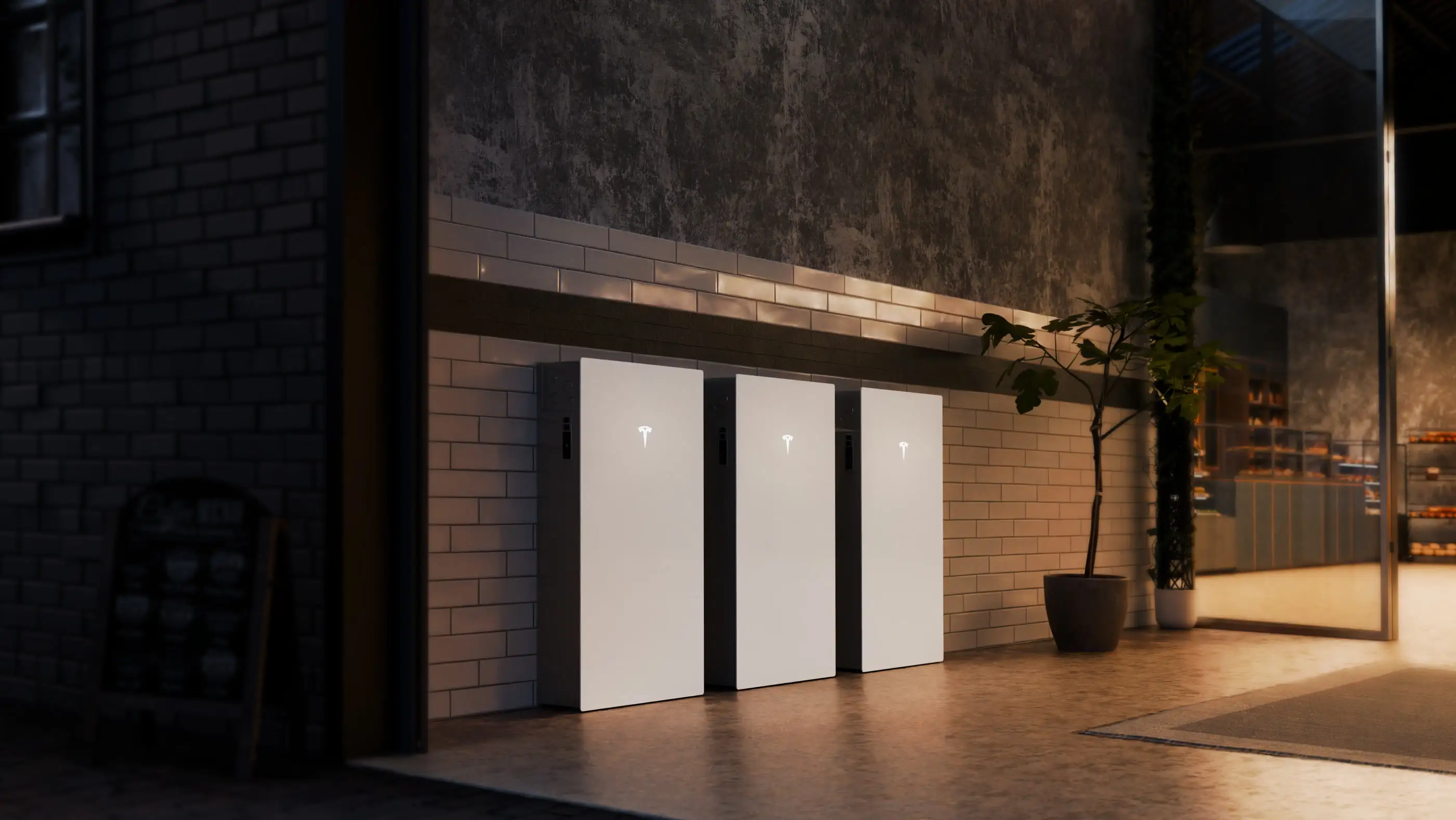 Three white Tesla home batteries standing side by side on a paved footpath against a white-tiled wall at night, under a timber awning with a potted fig beside them and a lit shop counter behind glass to the right