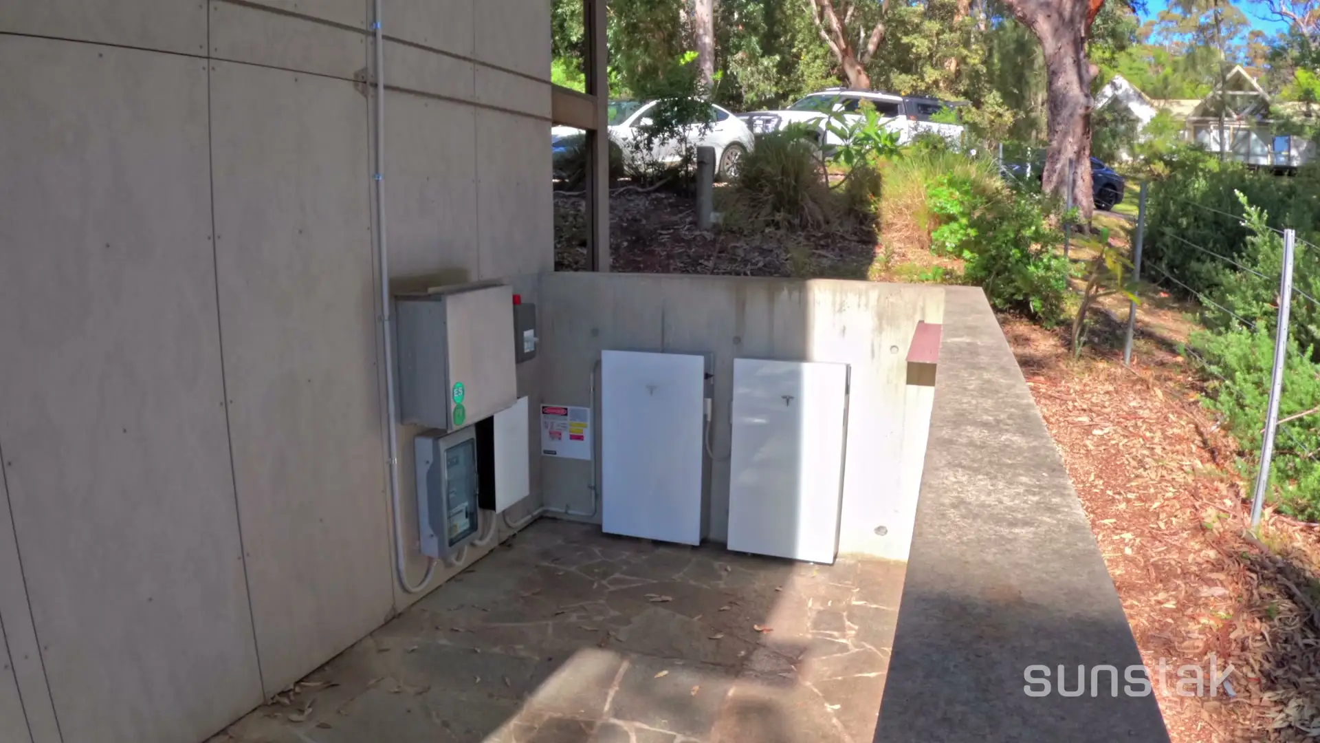 Two white Tesla-badged cabinets standing side by side against a concrete wall on a paved terrace outside a house, with a grey metal switchboard enclosure and a smaller meter box mounted on the timber wall to their left, bushland and parked cars behind