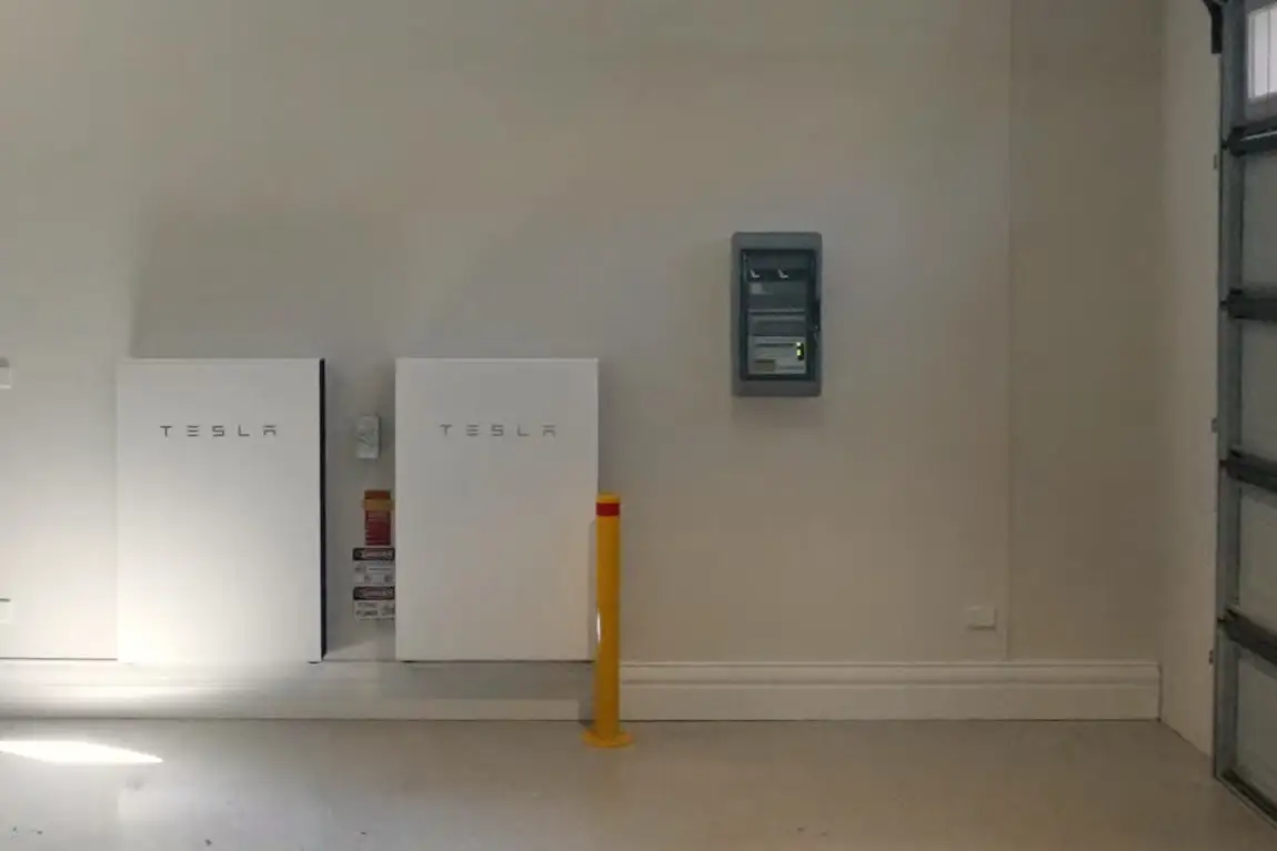 Two white Tesla-badged wall units mounted side by side on the rendered back wall of a garage, a grey switchboard enclosure on the same wall to their right, a short yellow bollard bolted to the floor in front of the right-hand unit, and the edge of a sectional garage door at the right of the frame