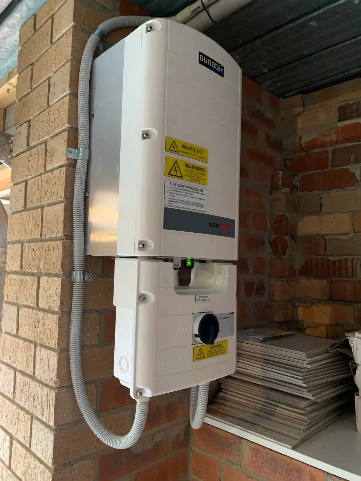 A Sunstak-badged SolarEdge inverter and its isolator mounted on a brick wall under a carport.