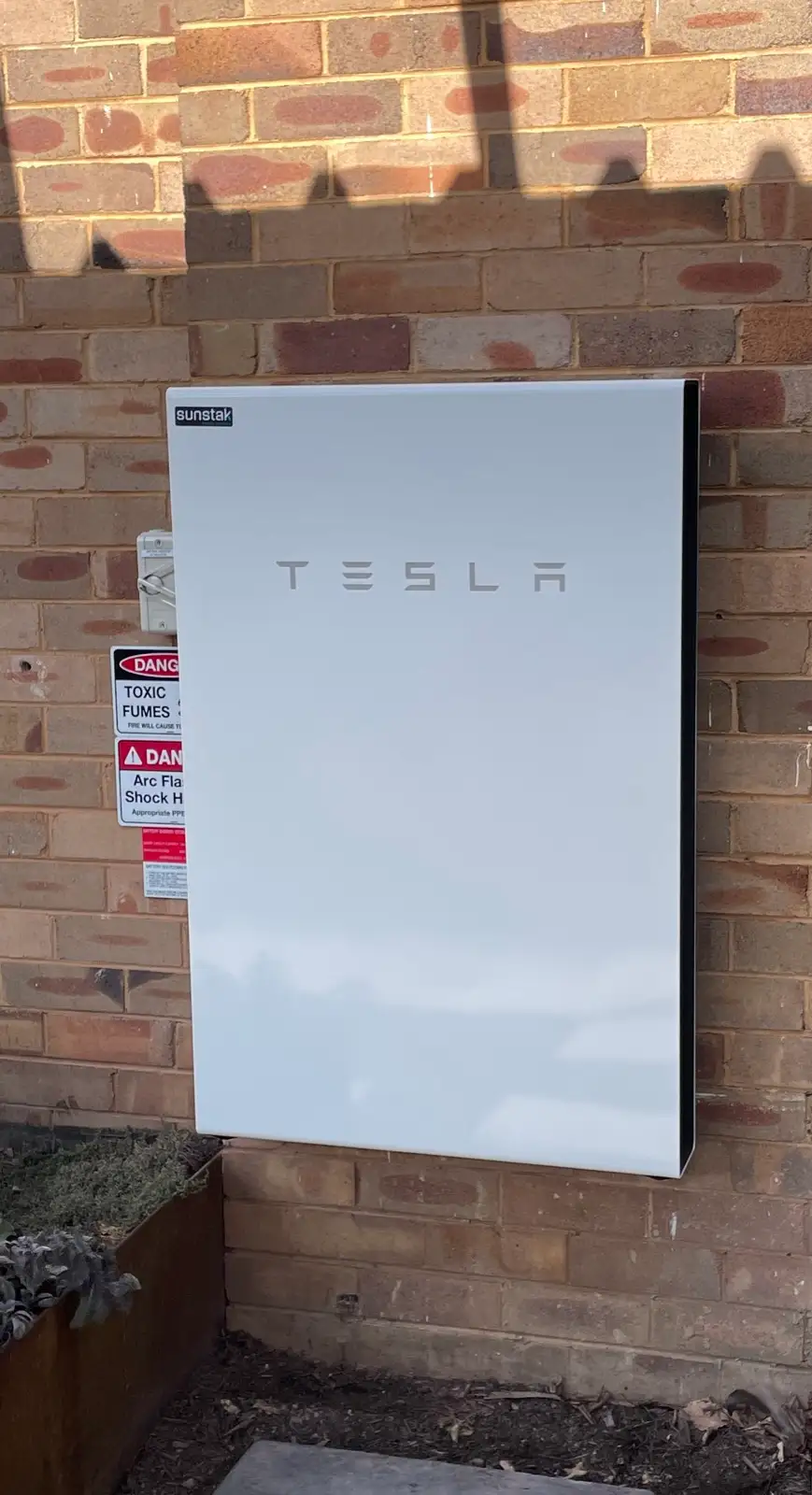A Tesla battery with a Sunstak badge on a red brick wall, its hazard placards to the left.
