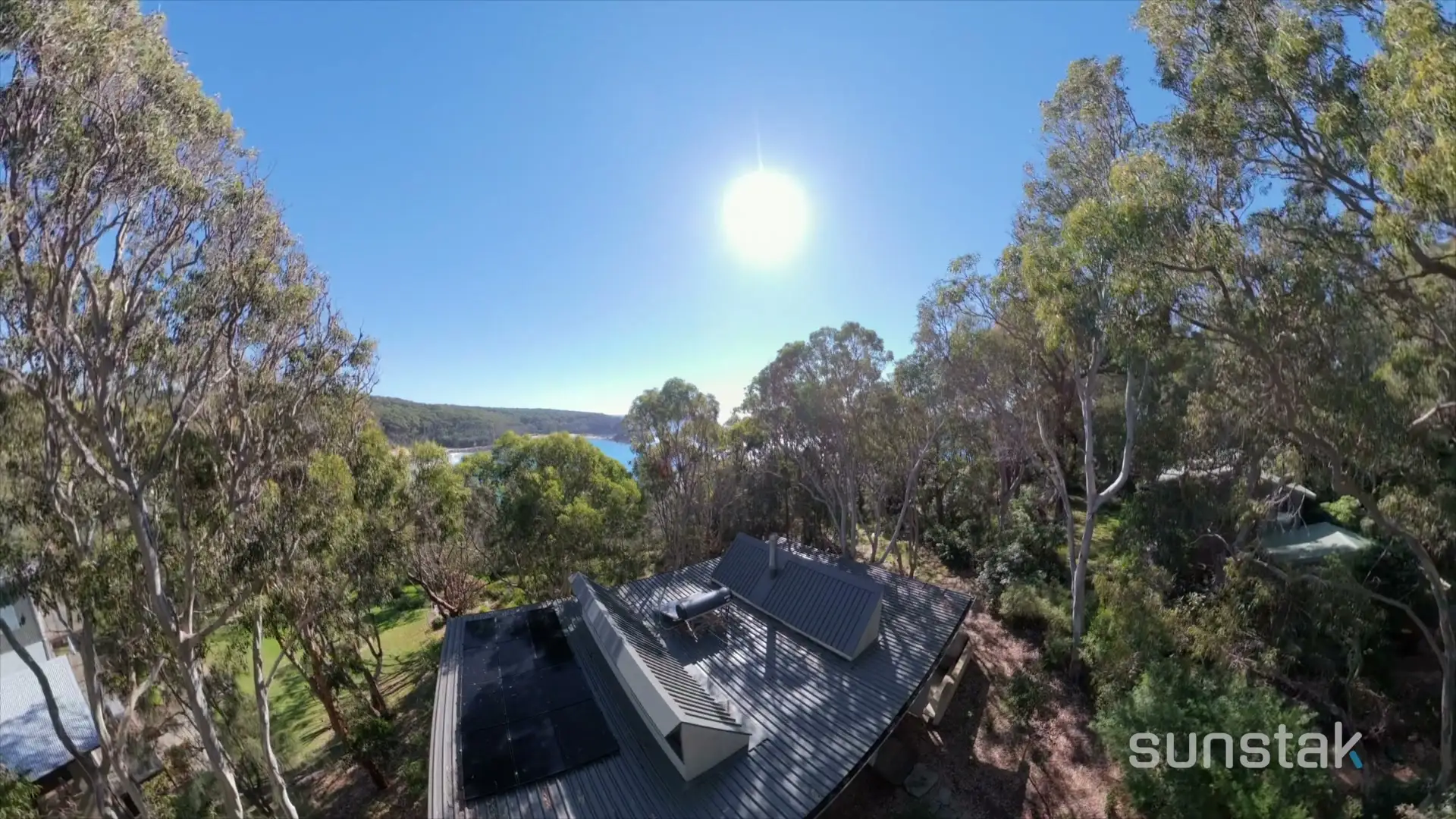 Seen from above, a house with a dark corrugated metal roof and a solar array on it, standing in a clearing among tall gum trees, with a beach and an inlet of blue water visible through the trees behind it