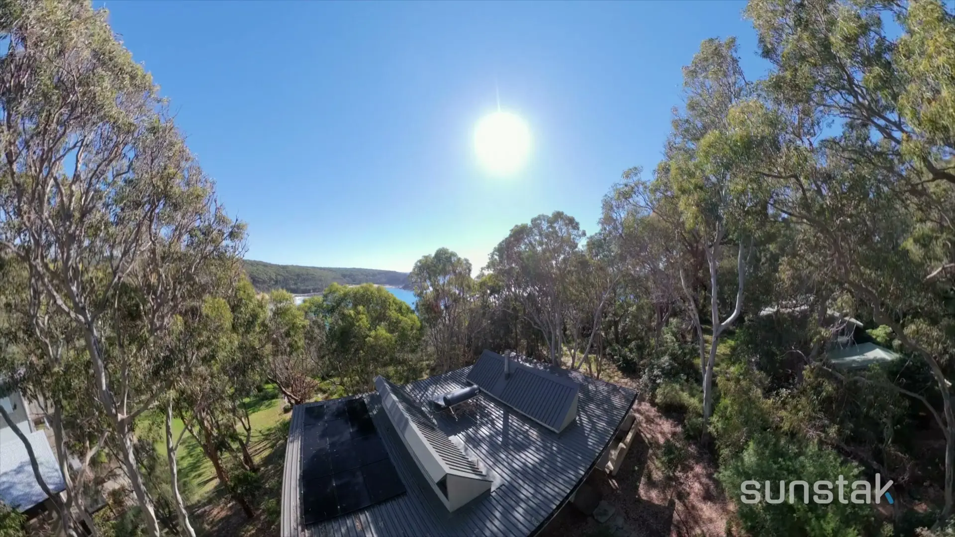 Seen from above, a house with a dark corrugated metal roof and a solar array on it, standing in a clearing among tall gum trees, with a beach and an inlet of blue water visible through the trees behind it and the sun high in a clear sky