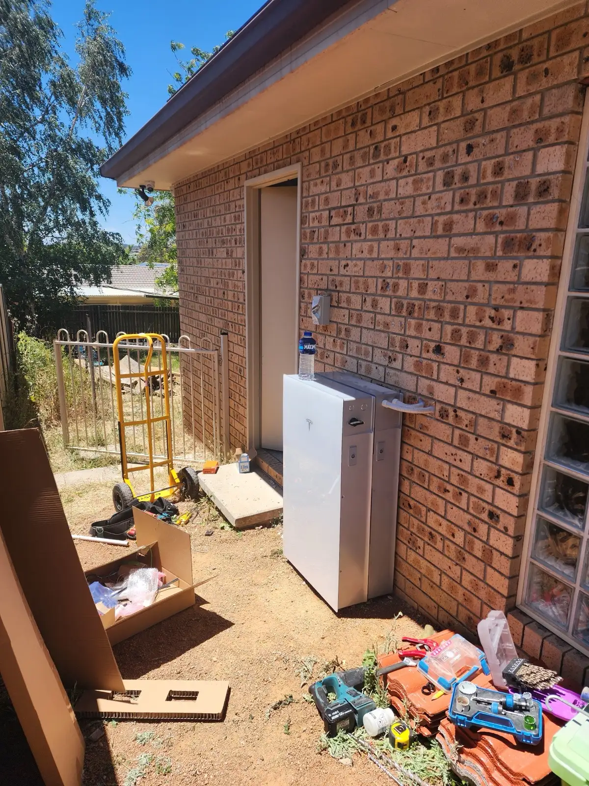 A Tesla battery and expansion unit standing against a brick wall on install day, tools and packaging still on the ground.