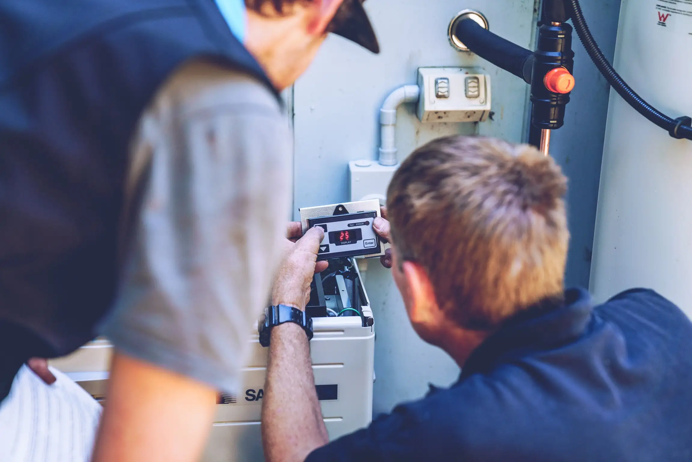 Two people crouched at an outdoor hot water unit, one holding a small controller with a red digital display reading 25 and the other leaning in beside them, with a weathered double power point, a black insulated pipe with an orange valve and the open cabinet of the unit below