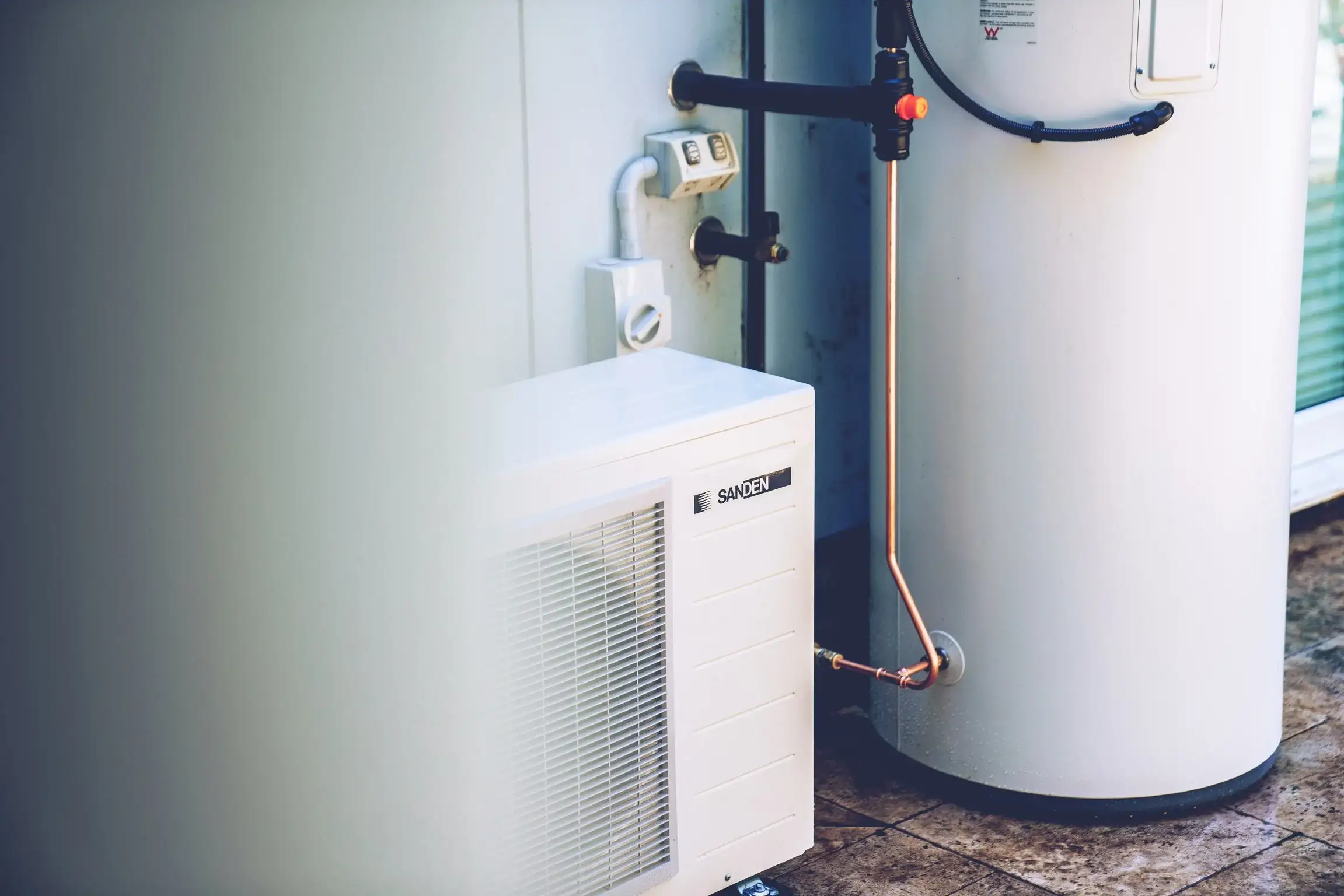 A cream Sanden outdoor heat-pump unit with a louvred grille standing on tiled paving beside a tall white storage tank, a bare copper pipe running between them, against a pale weatherboard wall with an isolator switch and a double power point above
