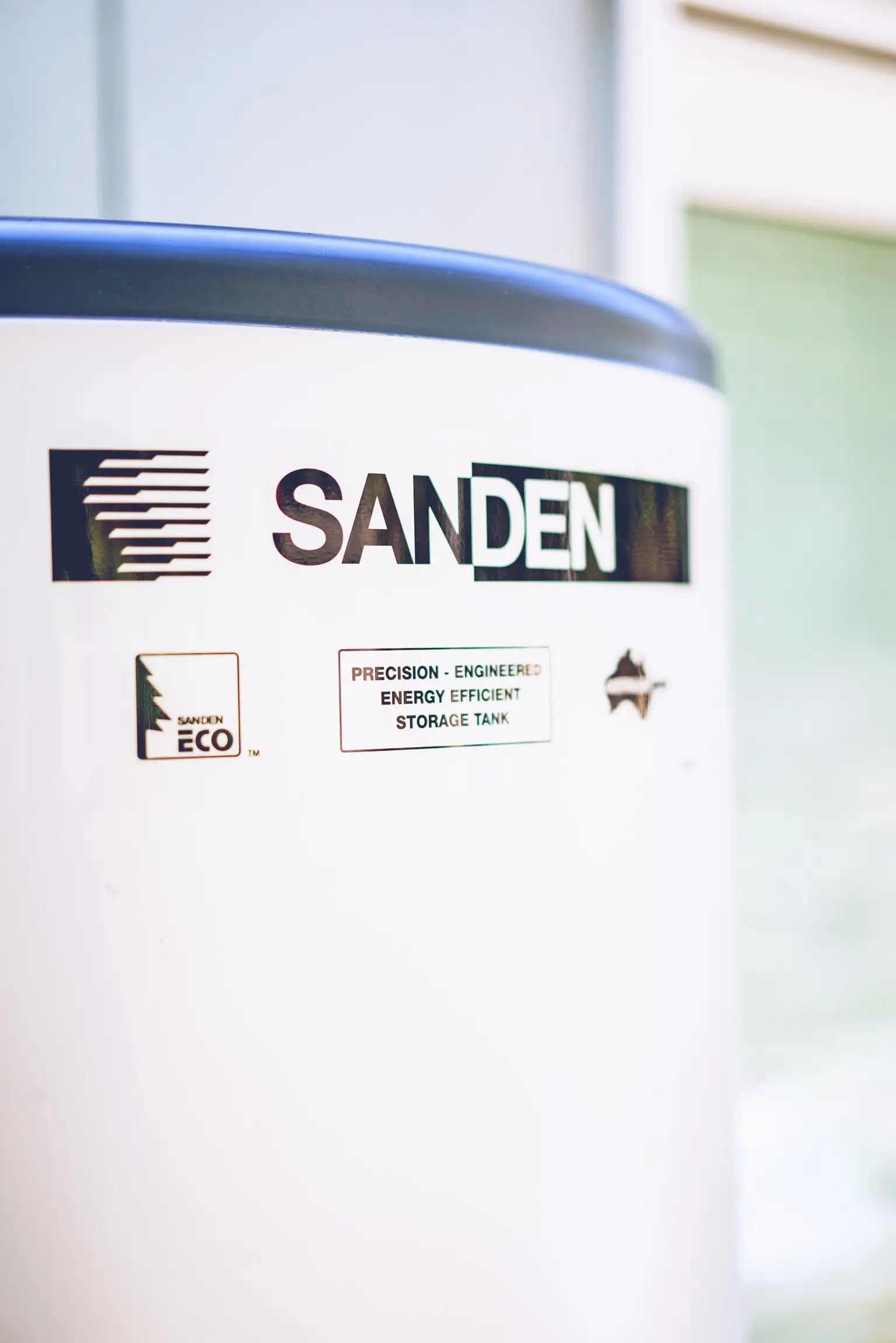 A close-up of the upper body of a white cylindrical storage tank with a dark grey top, carrying the SANDEN wordmark, a SANDEN ECO mark and a printed label reading PRECISION - ENGINEERED ENERGY EFFICIENT STORAGE TANK, with a pale wall and a green glazed door out of focus behind it