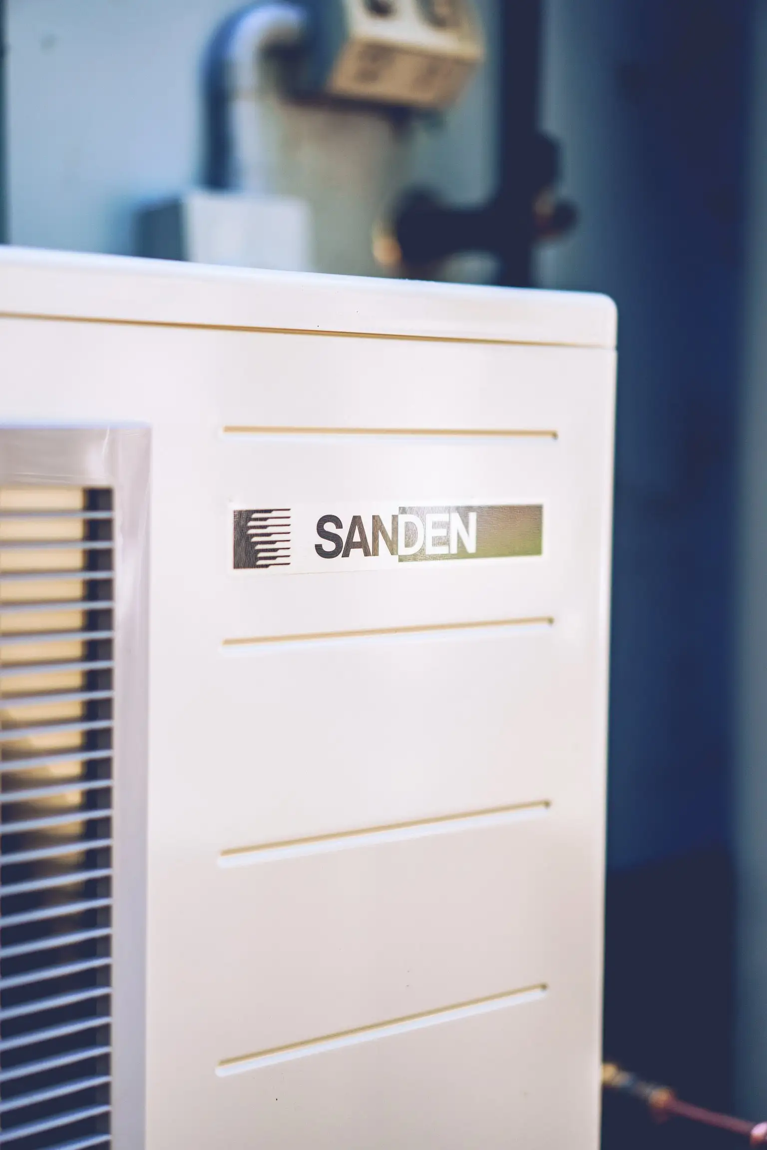 A close-up of the cream cabinet of an outdoor heat-pump unit, with the SANDEN wordmark on a foil label between four raised ribs and a tall louvred intake grille at the left edge, and a weathered double power point and black pipework out of focus behind it