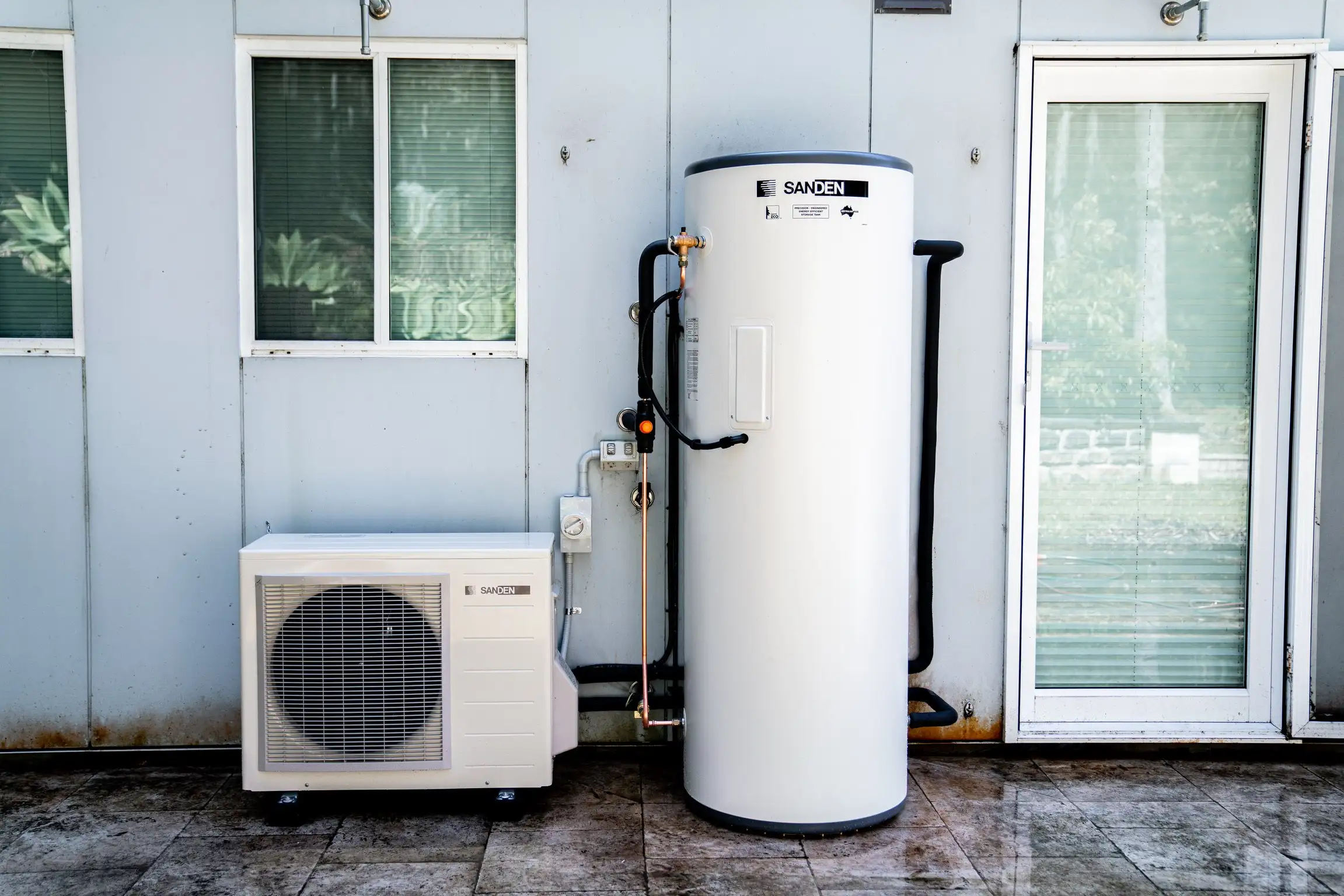 A cream Sanden outdoor heat-pump unit standing on wet paving beside a tall white Sanden storage tank, black and copper pipework running between them, against a pale panelled wall with a window to the left and a glazed door to the right