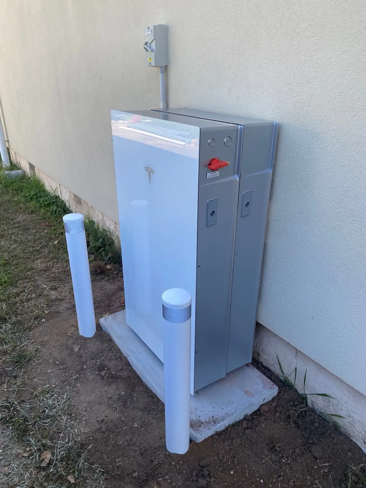 A Tesla battery and its expansion unit on a concrete pad against a rendered wall, bollards set in front.