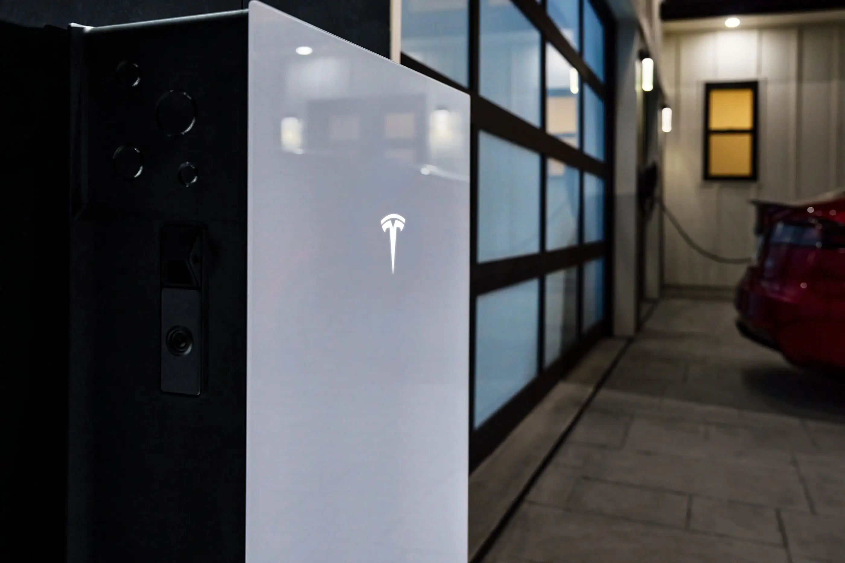 A white Tesla home battery seen close up at night, its dark grey side panel with four circular knockouts and a service handle turned towards the camera, with a glazed garage door, a small charger on the wall and the back of a red car in the corridor behind it