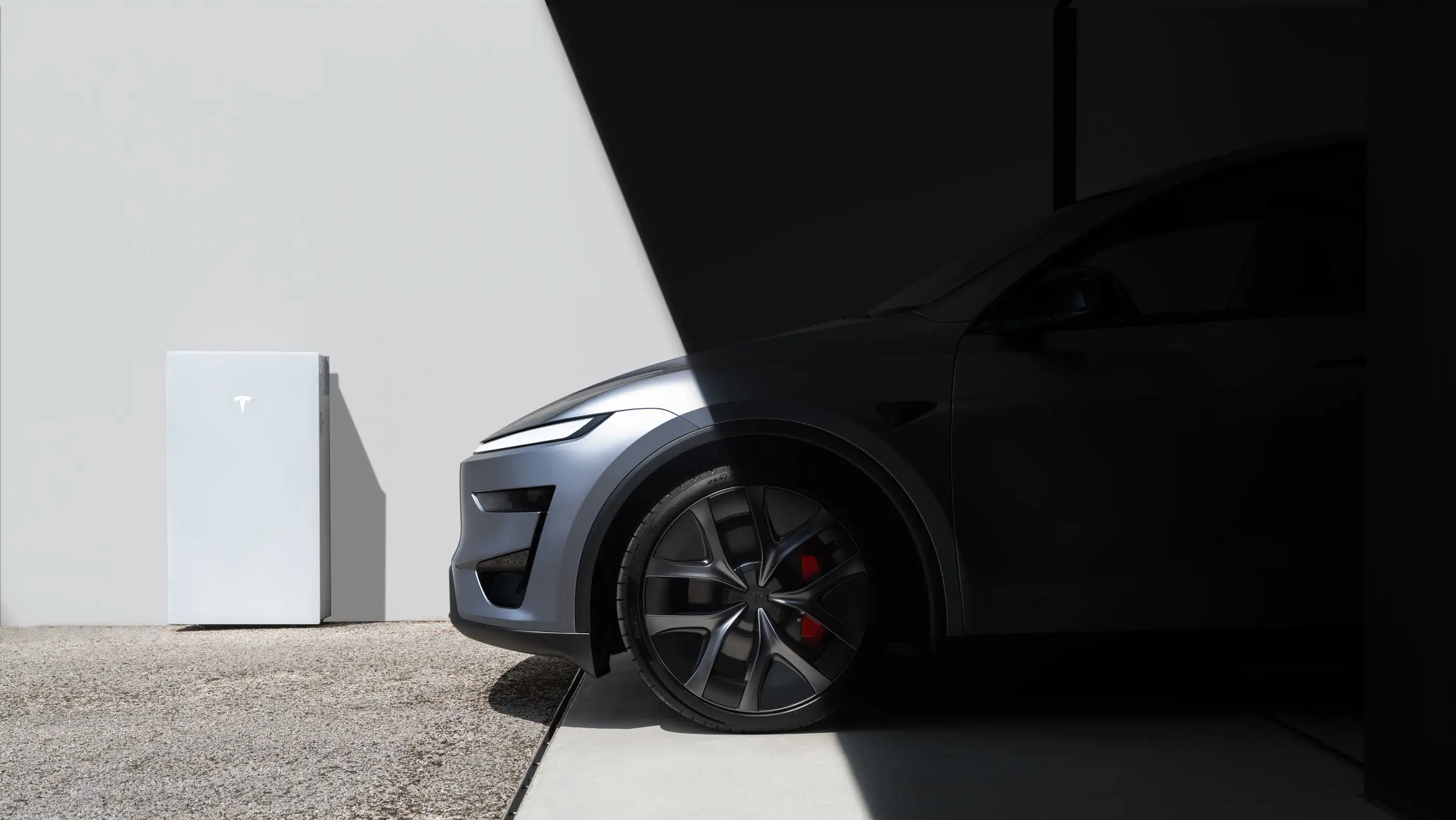 A white Tesla Powerwall standing against a pale rendered wall in hard sunlight, with a dark Tesla Model Y parked in deep shadow beside it and a gravel strip along the edge of the driveway