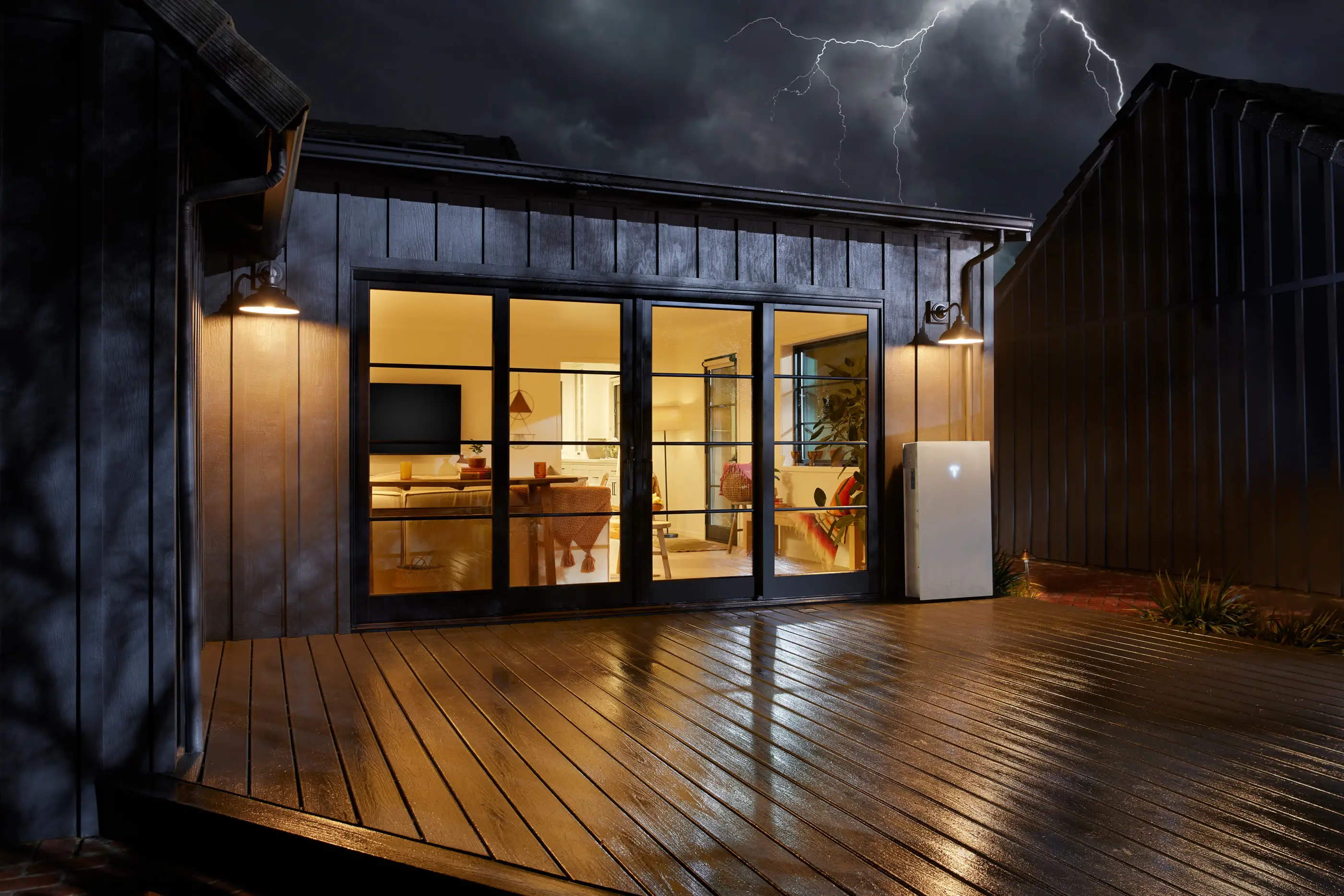 Lightning over a dark timber house at night, its wet deck reflecting the warm light from a lit kitchen behind glass doors, with a white home battery on the wall beside them