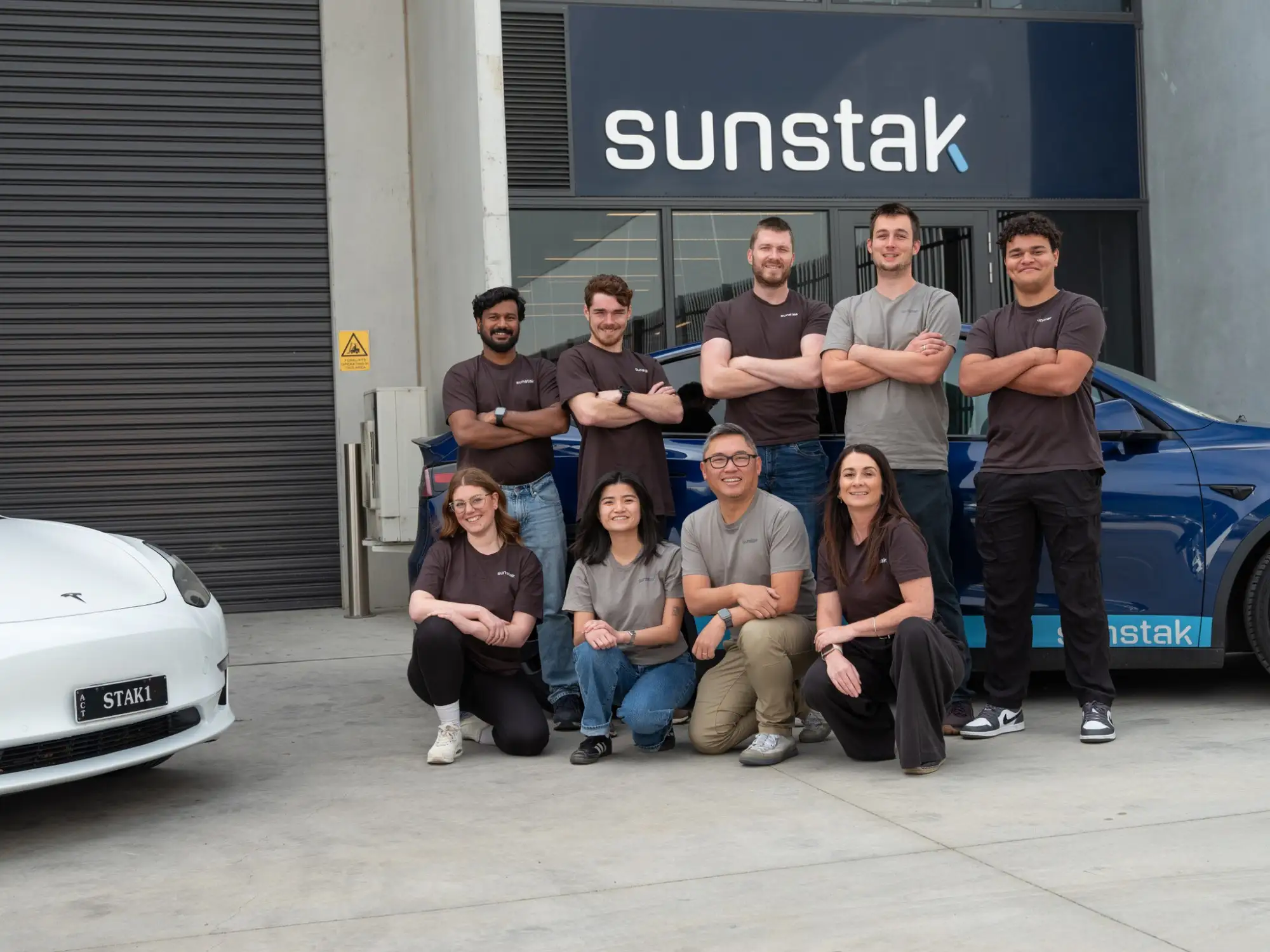 Nine people in dark and grey Sunstak t-shirts outside a warehouse, four kneeling in front and five standing behind, under a lit sunstak sign above the glass doors, with a white car at the left and a blue car carrying sunstak lettering along its side at the right