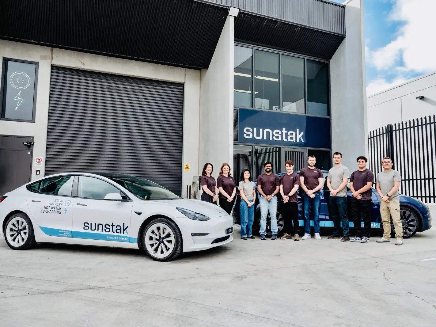 Nine people standing in a line on concrete outside a pale industrial building with a dark sign reading sunstak above a roller door, in front of a dark blue car with a pale blue lower-body wrap; a white car lettered sunstak is parked across the foreground