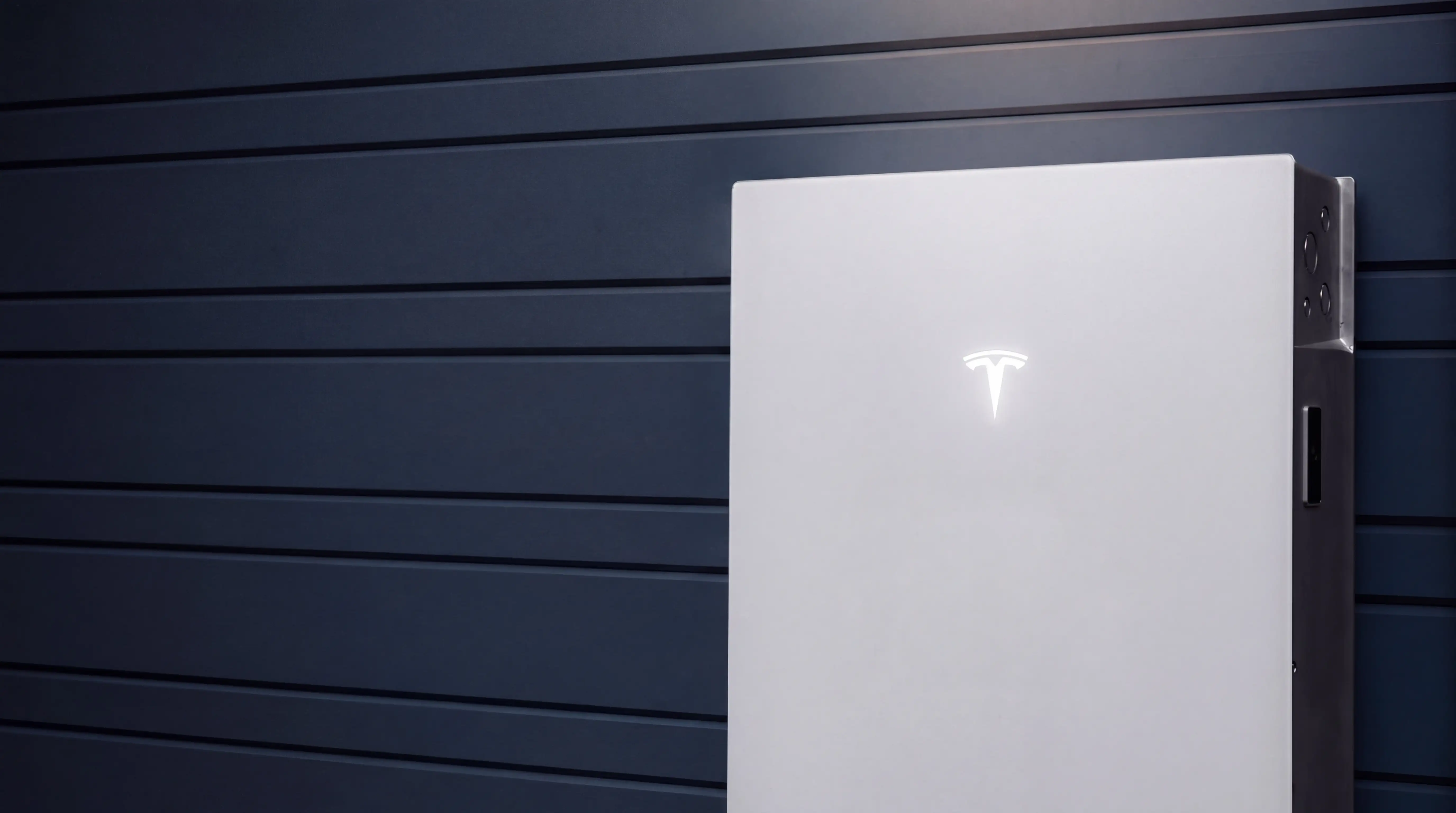A white home battery carrying a lit Tesla mark, mounted on the dark blue horizontal cladding of a house wall, with its dark grey side rail, vent knockouts and service handle catching the light along the right-hand edge