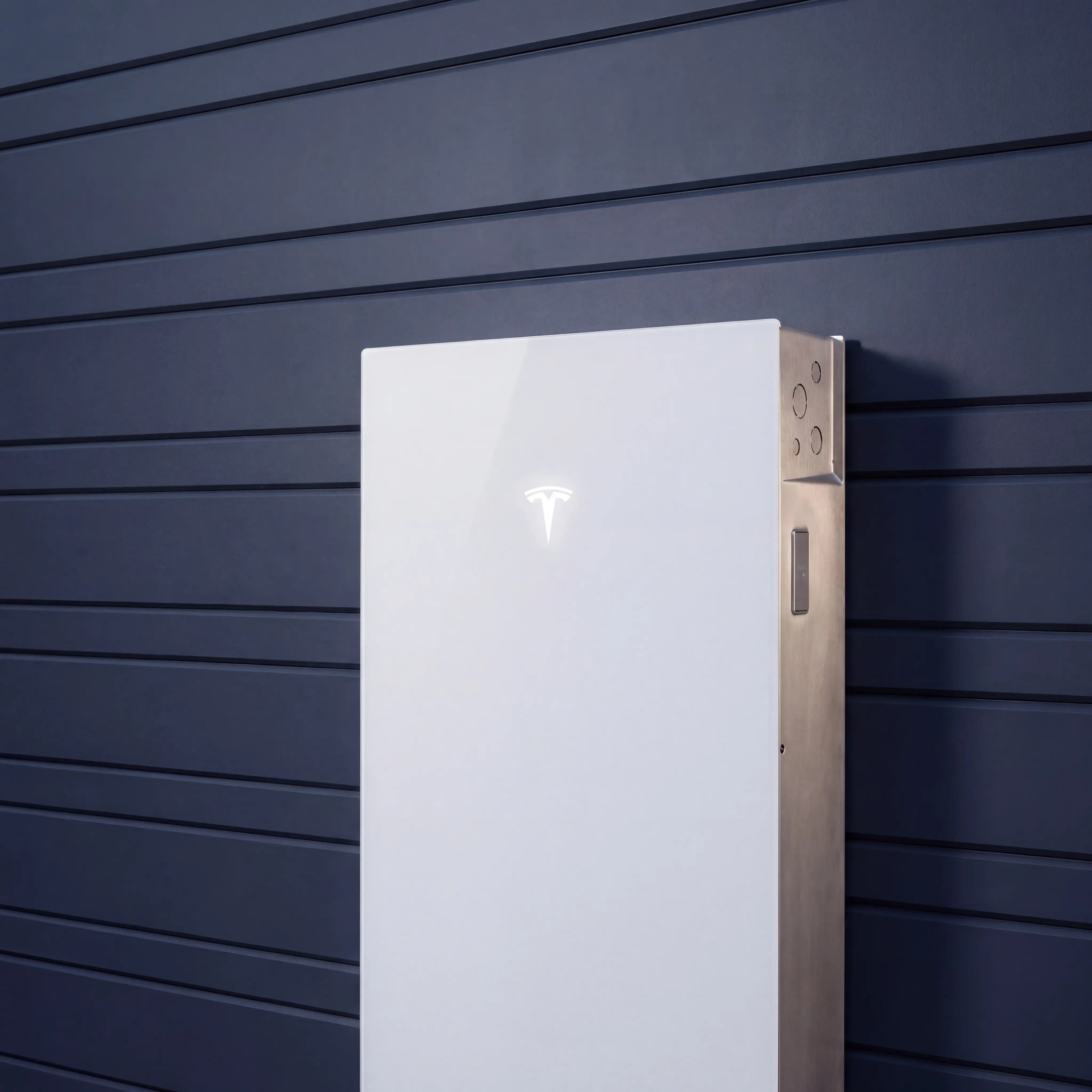 A white Tesla Powerwall 3 standing against the blue-grey weatherboard wall of a house at dusk