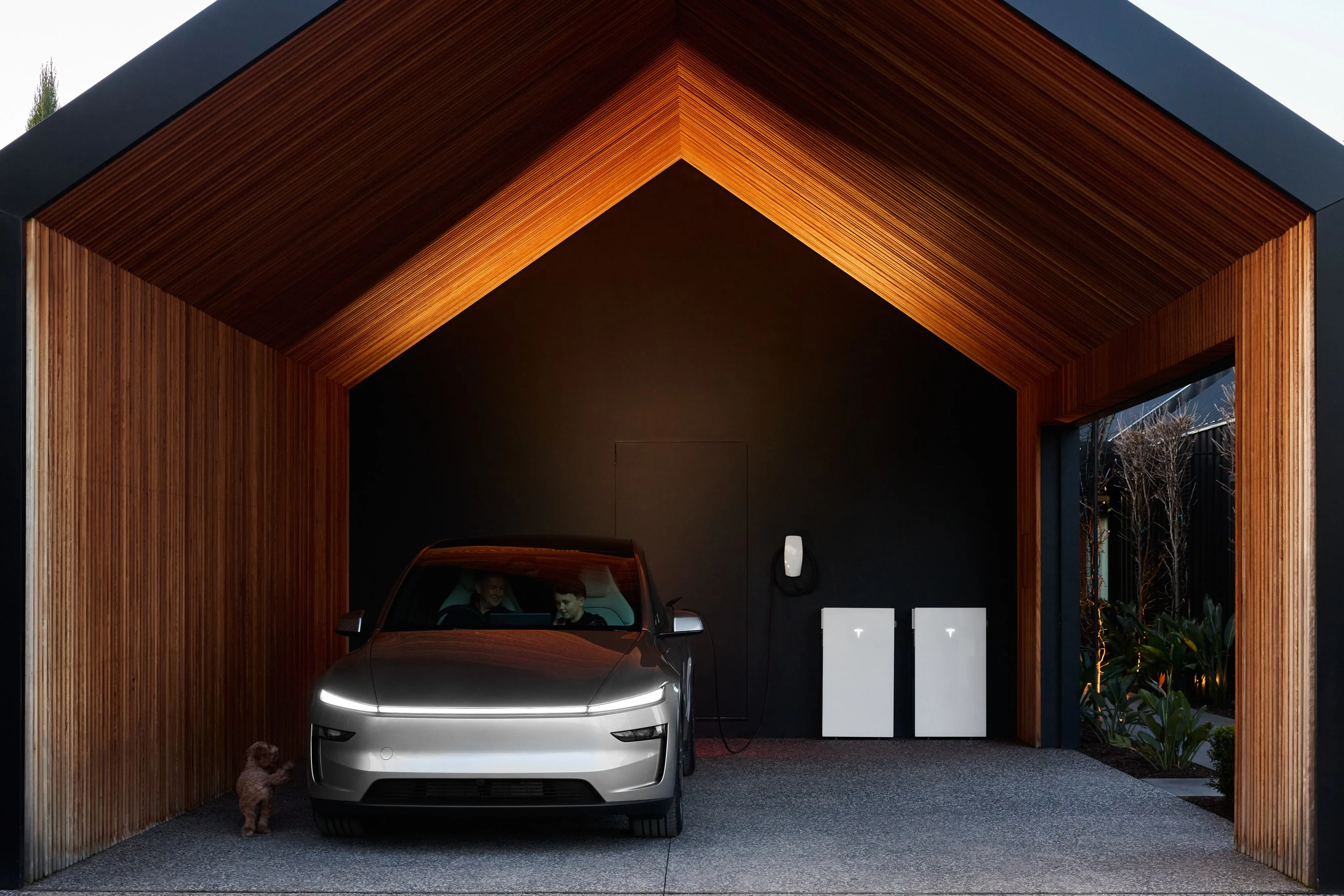 Two white Tesla Powerwall 3P units with lit Tesla marks on the back wall of a timber-lined garage, a silver car parked in front