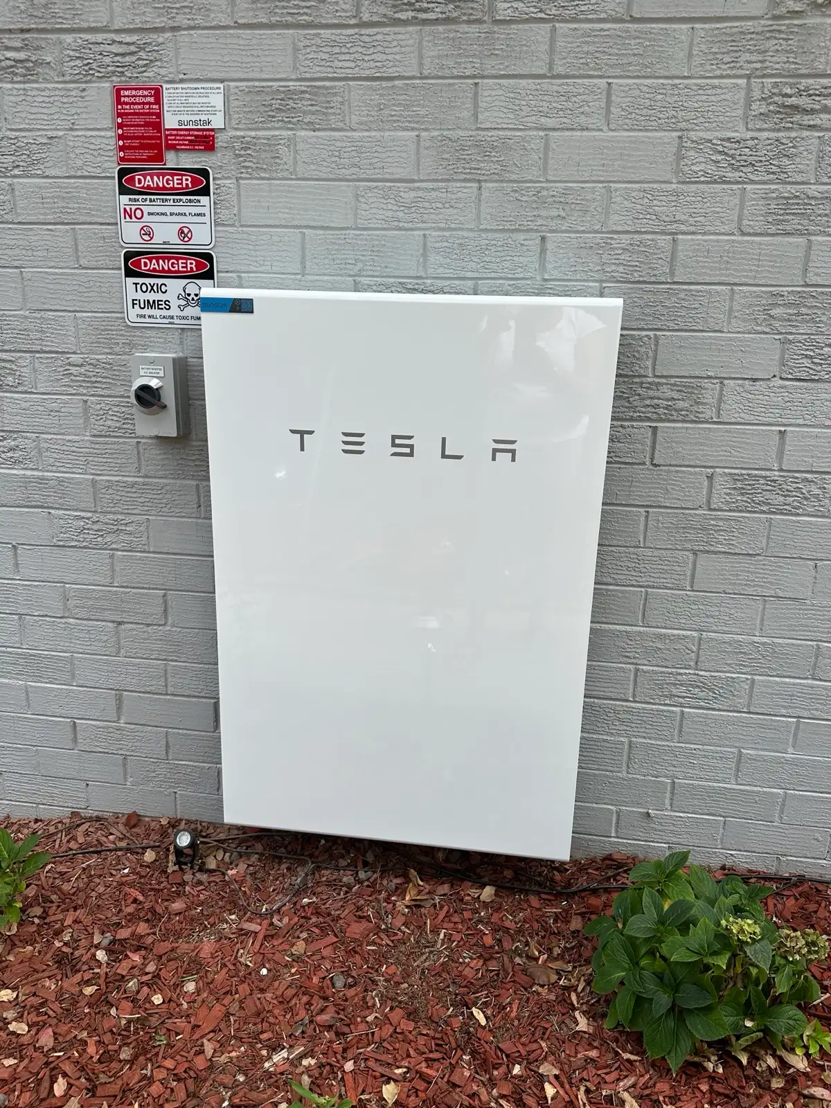 A Tesla battery on a painted brick wall above a mulched garden bed, shutdown placards to the left.