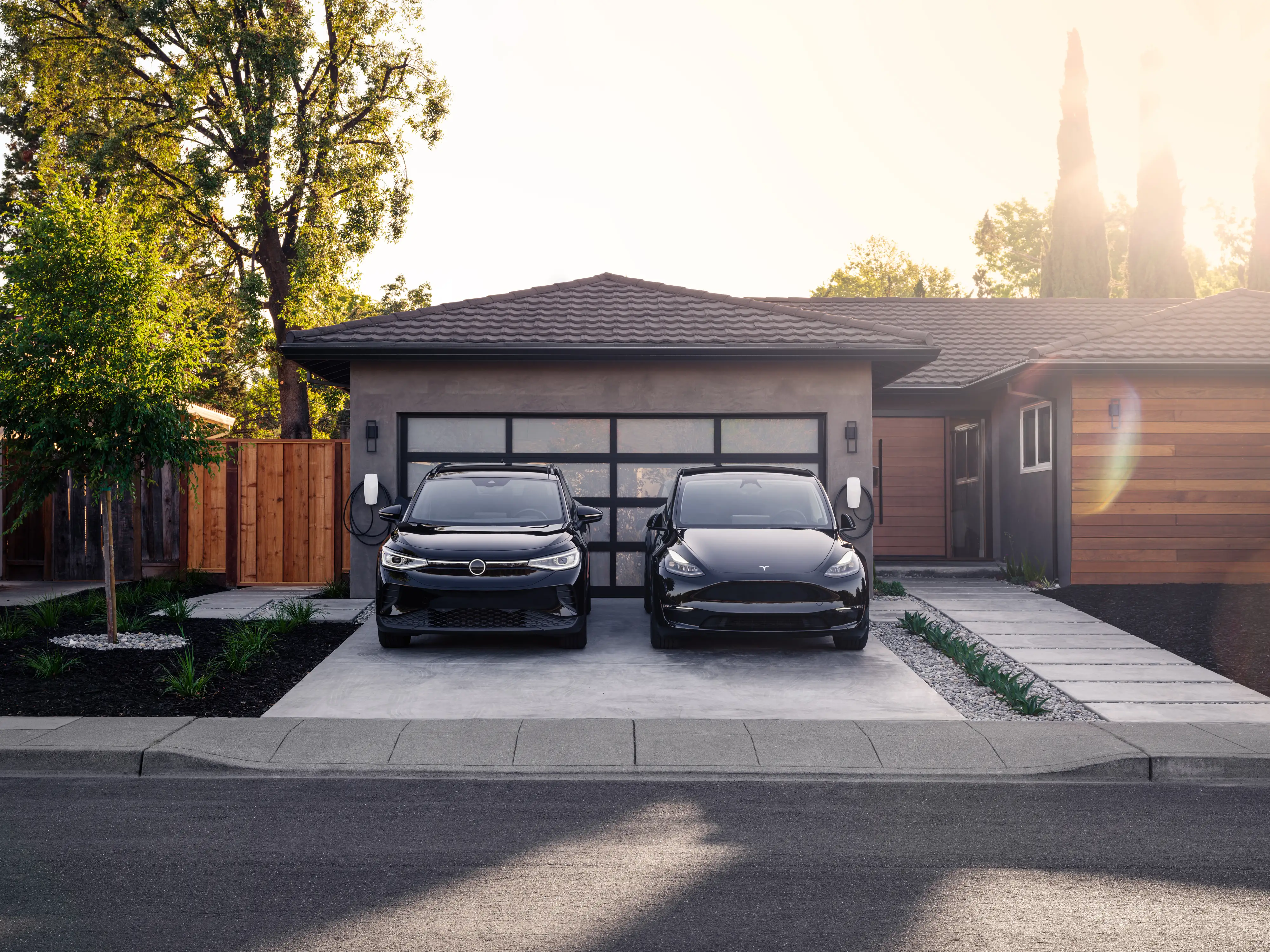 Two black electric SUVs parked nose-on side by side in a driveway at sunset, each plugged into its own white Tesla Wall Connector mounted either side of a glazed garage door. The car on the right carries a Tesla badge; the one on the left is another make