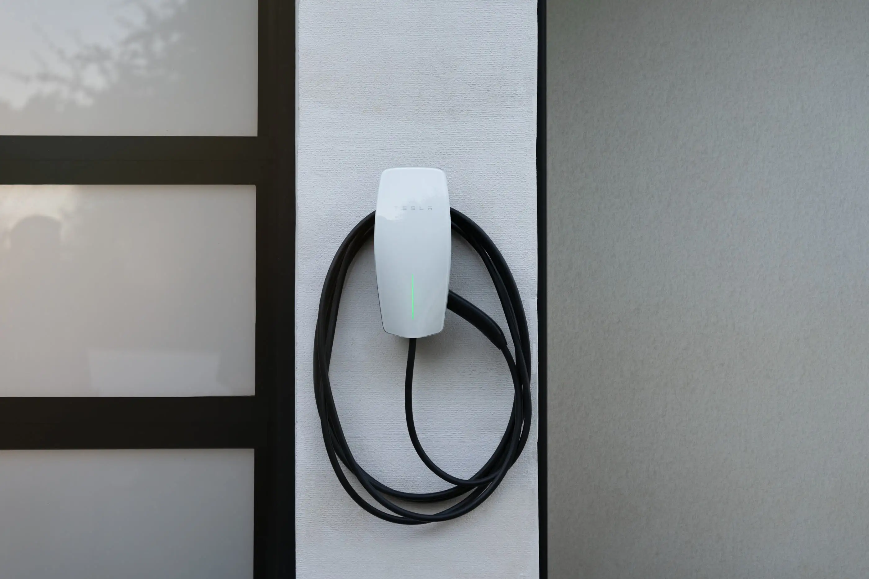 A white Tesla Wall Connector mounted on a narrow white rendered pier between two dark door frames, its black cable coiled below it and a green light showing on the faceplate