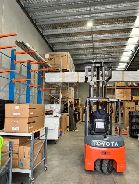 A person driving an orange forklift down the aisle of a warehouse, between racking holding stacked flat panels and pallets of cartons, under a steel roof