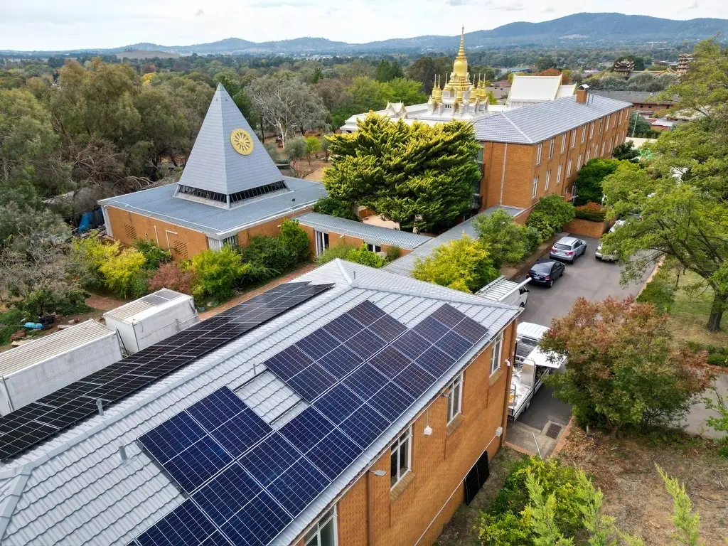 Aerial view of solar panels across the tiled roof of a long brick building, with a spire-roofed hall beside it and gilded temple roofs among trees behind