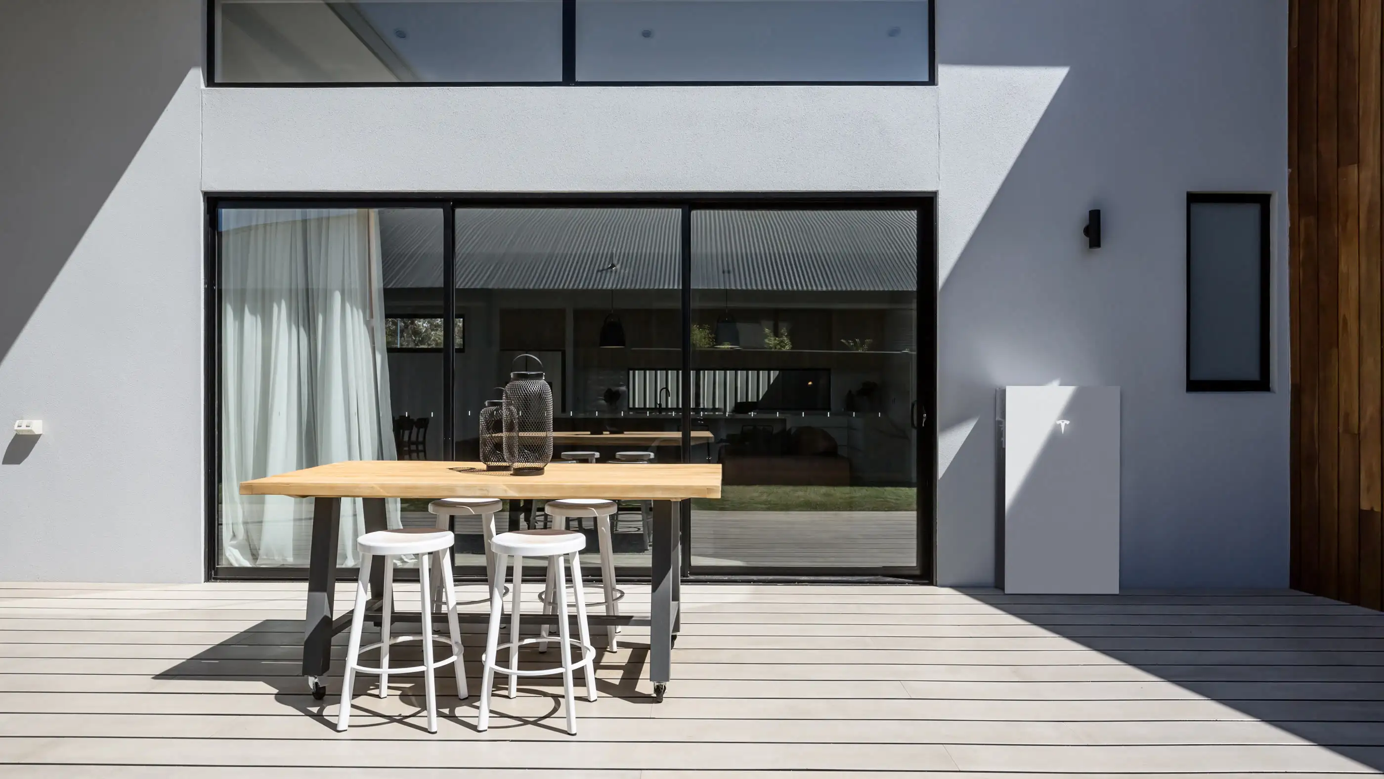 A white Tesla Powerwall 3 mounted on a white rendered wall beside glass sliding doors and a timber deck, in daylight