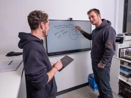 Two people in hoodies standing at a wall-mounted screen in an office, one of them reaching up to the screen, which carries a hand-drawn sketch and a few lines of working; the other is holding a tablet