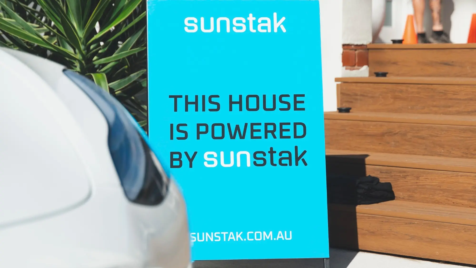 A cyan sign board standing on a driveway, lettered sunstak above the words THIS HOUSE IS POWERED BY SUNSTAK with the address sunstak.com.au along the bottom, the front of a white car in the left foreground and timber steps with two orange traffic cones behind it